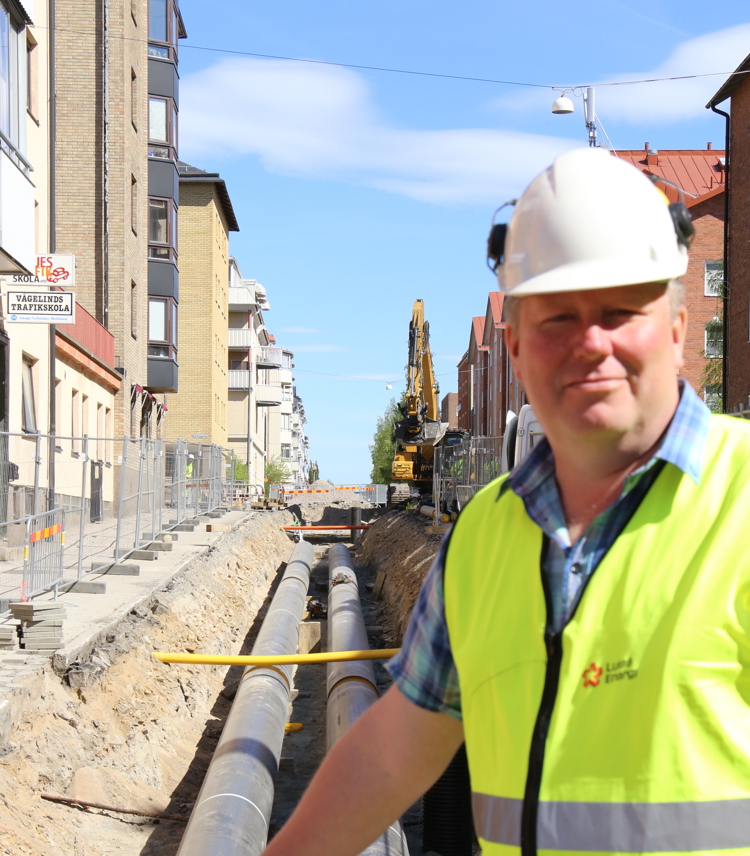 Äldre man med varselväst och hjälm som står framför en uppgrävd väg med fjärrvärmeledningar